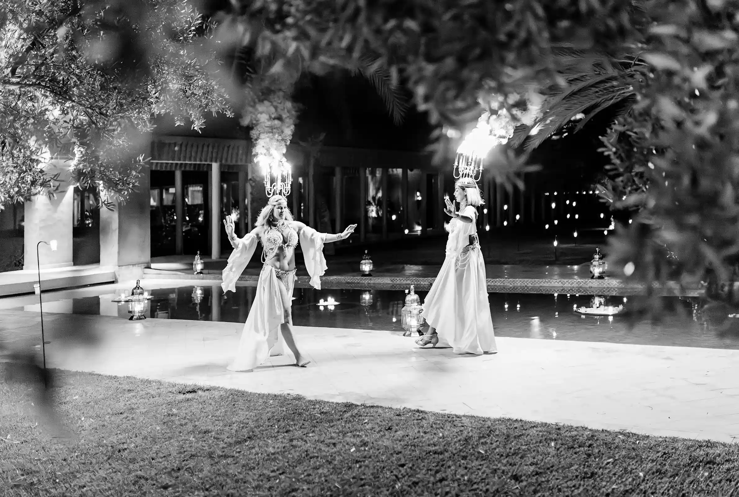 Deux danseuses portant des chandeliers allumés au bord d’une piscine lors d’un mariage à Marrakech, en noir et blanc. Two dancers balancing lit candelabras beside a swimming pool during a wedding in Marrakech, photographed in black and white.